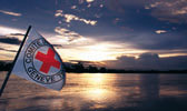 Foto: Eine Flagge des Roten Kreuzes weht vor der Weite des Horizontes. Die untergehende Sonne spiegelt sich auf der Wasseroberfläche. Dieses Foto ist gleichzeitig ein Link und führt Sie zu der Unterseite: Grundsätze.