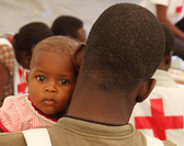 Foto: Ein haitianisches Baby schaut über die Schultern eines  Rotkreuzhelfers.